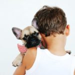 Shows a Pug being held by a boy. the boys back is facing the camera and the Pugs little head is on his shoulder